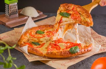Fresh delicious fast food. A piece of hot pizza with melting cheese on the table. Traditional Margherita pizza with tomatoes, basil and mozzarella cheese close-up on a dark background