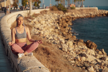 Fototapeta na wymiar Adult pretty lady sitting by the seashore in lotus position in sports clothes on sunny day