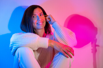 Smiling young brunette woman wearing a knitted sweater and white pants lit with a blue spotlight and a pink spotlight