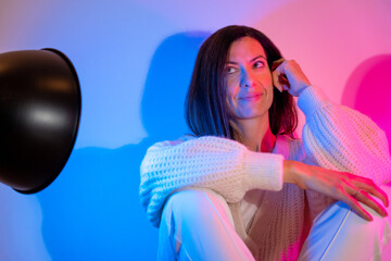 Smiling young brunette woman wearing a knitted sweater and white pants lit with a blue spotlight and a pink spotlight