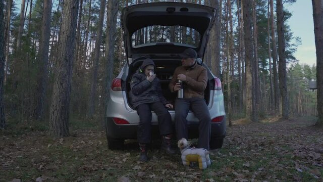 Teenage boy with father seated on open trunk of suv standing in forest and drinks hot tea. Traveling and adventure conception.