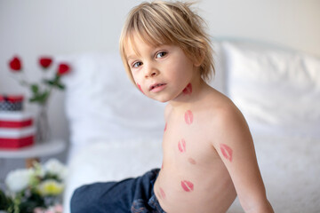 Beautiful blond toddler child, boy with lipstick kisses on his body, holding red rose for Valentine