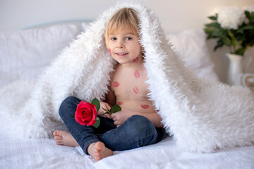 Obraz premium Beautiful blond toddler child, boy with lipstick kisses on his body, holding red rose for Valentine