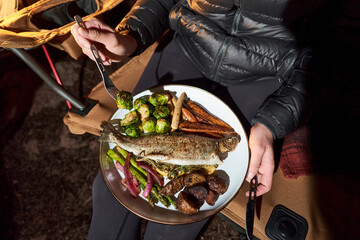 Woman has camp dinner with trout, brussel sprouts and carrots.