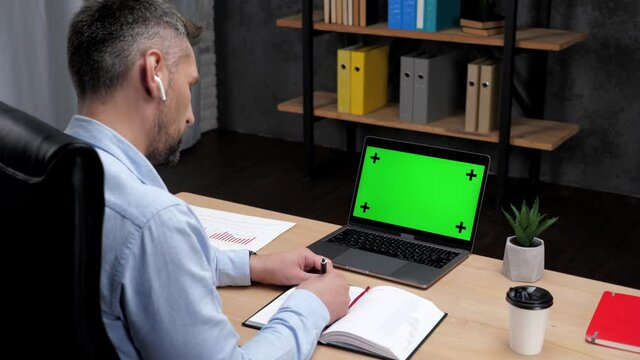Man professional stock trader broker makes notes in notebook uses laptop with green screen. Businessman writes in notebook looks mock up chroma key computer display sitting on chair at desk in office