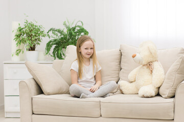 Photo of little girl in white clothes sitting on white sofa with teddy bear.
