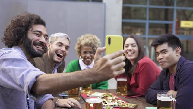 Group happy diverse friends doing online videocall selfie in outdoors bar to share celebration on social media