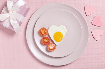 Fried eggs in the shape of a heart on a pink plate with tomatoes and gift wrapping on a pink background.