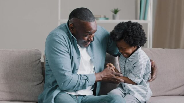 Funny mature african american father with little daughter cuddling sitting on sofa in room playing fun embrace loving dad tickles kid girl play game together child smiling laughing happy family hugs