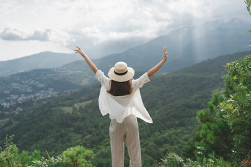 Young beautiful woman with long hair stands with her back raising her hands up, a beautiful view of the mountains and the forest. Travel and healthy lifestyle.