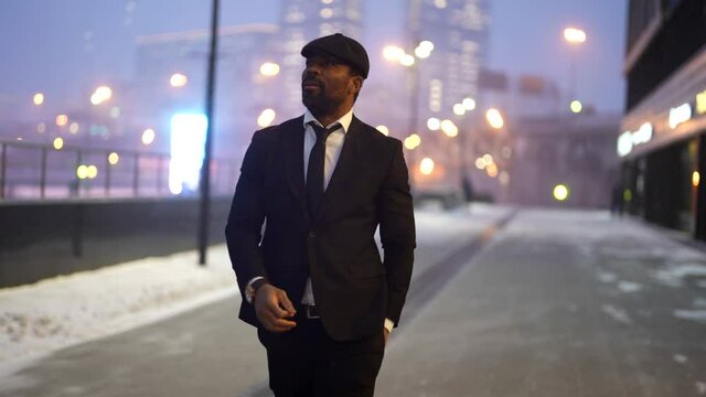 successful stylish black man is strolling on city street in winter evening, night city life