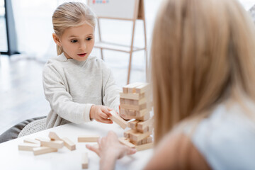 Girl holding wooden block while playing with blurred mother at home.