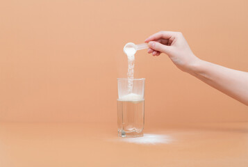 Woman pours collagen powder or protein in a glass of water on a beige background. A healthy and anti aging supplement.