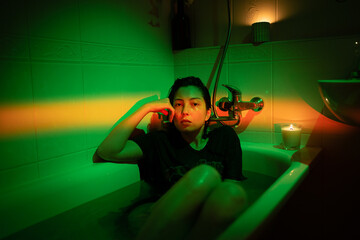 Portrait of unhappy woman taking bath dressed and tired. Drunk young female lying in cold water in bathroom. Anxious girl in t-shirt hiding from problems. Colorful neon light. Midlife crisis concept
