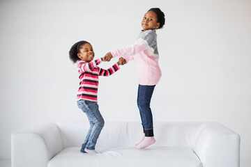 Pretty african american children holding hands and jumping together on white cozy sofa. Two happy sisters with curly hair enjoying carefree lifestyles during childhood.