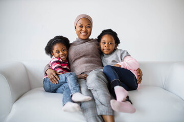 Beautiful african american islamic woman in headscarf sitting on couch with her two pretty daughters and smiling on camera. Sisters embracing mother in studio.