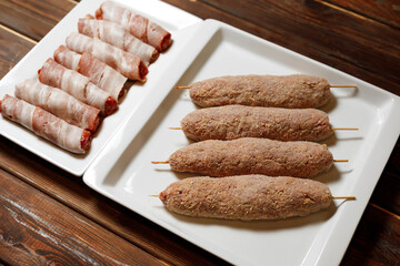 There are four sausages with sticks in them and a few rolls of meat in white plates on the wood table.