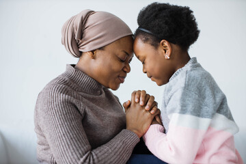 African american islamic mother in hijab and her little daughter sitting face to face, holding hands and keeping eyes closed. Studio with white background.