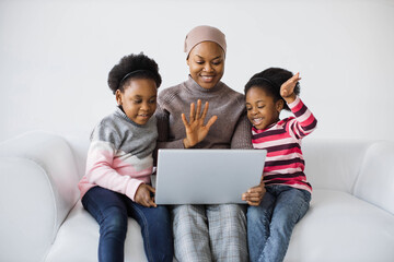 Beautiful islamic mother and her two pretty daughters using modern laptop for video call in studio. African american family of three sitting on couch, waving hands and smiling.