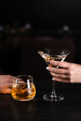Two cocktails in hand on a dark background. A man's hand and woman's hand holds a glass of cocktails on bar counter