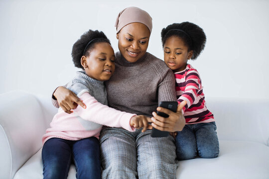 Two cute sisters and their caring mother using modern smartphone for taking selfie while resting on comfy couch. African american family having fun while spending time together .