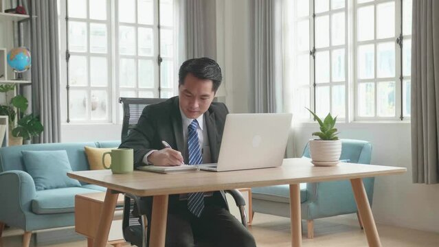 Asian Businessman In Business Suit Looking At Computer Screen And Writing In Notebook On A Table While Working At Home. 
