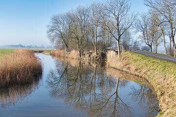 Bare trees reflect in the smooth water of a watercourse through the meadows of the Dutch province...