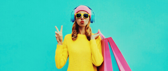 Fashionable portrait of stylish young woman listening to music in headphones with shopping bags posing wearing a yellow knitted sweater, pink hat on blue background - obrazy, fototapety, plakaty