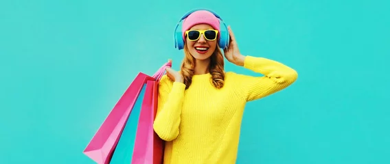 Photo sur Aluminium Magasin de musique Portrait coloré d& 39 élégante jeune femme souriante écoutant de la musique dans des écouteurs avec des sacs à provisions posant portant un pull en tricot jaune, un chapeau rose sur fond bleu