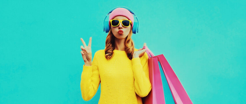 Fashionable portrait of stylish young woman listening to music in headphones with shopping bags posing wearing a yellow knitted sweater, pink hat on blue background