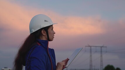 Civil engineer, woman specializing in power supply, works outdoors. Concept of green energy, ecology. Modern technologies. Power engineer in a protective helmet checks the power line, digital tablet