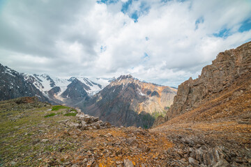 Obraz premium Vivid mountain landscape near precipice edge with sharp stones with view to sunlit large pinnacle with snow under cloudy sky. Picturesque view from stone hill to mountain peak with shadows from clouds
