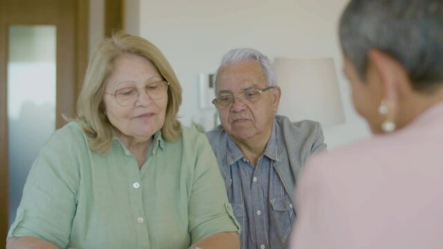 Mature couple negotiating real estate agreement with realtor. Front view of happy Caucasian wife and husband in eyeglasses preparing for signing contract, making deal. Real estate purchase concept
