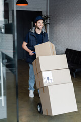 Positive courier standing near cart with packages in hallway.