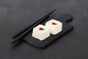 Festive creamy white chocolate mousse cakes with strawberry jelly heart, in the form of square sweet sushi, sprinkled with coconut chips, on a serving board on a dark gray background. Valentine's Day