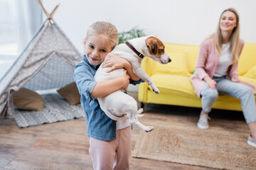 Happy kid hugging jack russell terrier near blurred mom at home. © LIGHTFIELD STUDIOS