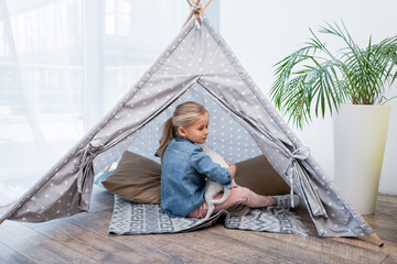 Girl hugging jack russell terrier in teepee near plant at home. © LIGHTFIELD STUDIOS