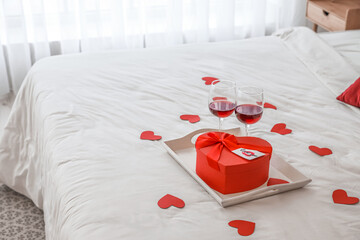 Tray with gift for Valentine's Day and glasses of red wine on bed
