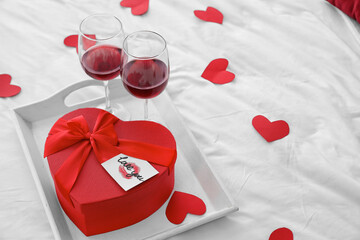 Tray with gift for Valentine's Day and glasses of red wine on bed