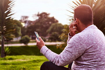 sentado al lado del camino de un pueblo esta un joven contestando una llamada a su celular con sus audífonos inalámbricos 