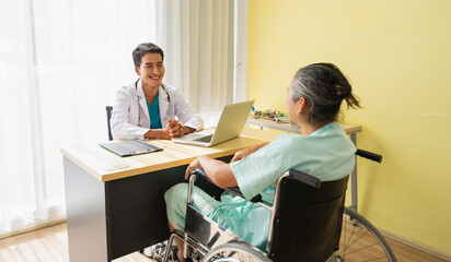 Doctor smile and happy sitting with laptop looking to the elderly patient man sitting on wheelchair. Doctor or nurse take care of senior patient health visiting at hospital or clinic