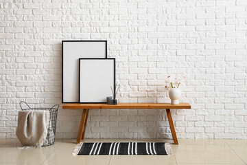 Wooden bench with blank frames, reed diffuser, flowers in vase and basket near white brick wall