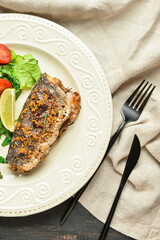 Plate with delicious sea bass fish, vegetables and cutlery on dark wooden background, closeup