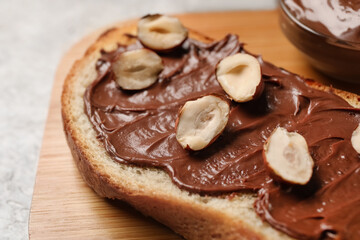 Fototapeta na wymiar Wooden board of bread with chocolate paste and hazelnuts on light background, closeup