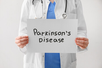 Female doctor holding paper sheet with text PARKINSON'S DISEASE on light background, closeup