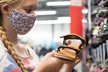 New normal retail shopping. Teenage blond girl wearing face mask choosing walking shoes sandalds at...