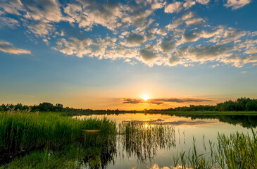 Fototapeta na wymiar Amazing view at scenic landscape on a beautiful lake and colorful sunset with reflection on water surface among green reeds and glow on a background, spring season landscape