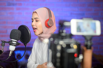 Young smiling muslim female singer wearing headphones with a microphone while recording song in a...