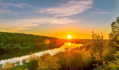 Scenic view at beautiful spring sunset on a shiny river valley with green branches, trees, bushes, grass, golden sun, calm water ,deep blue cloudy sky and forest on a background, spring landscape
