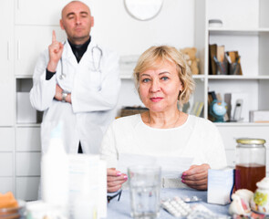 Mature woman calls a doctor at home, sitting at a table with medicines.